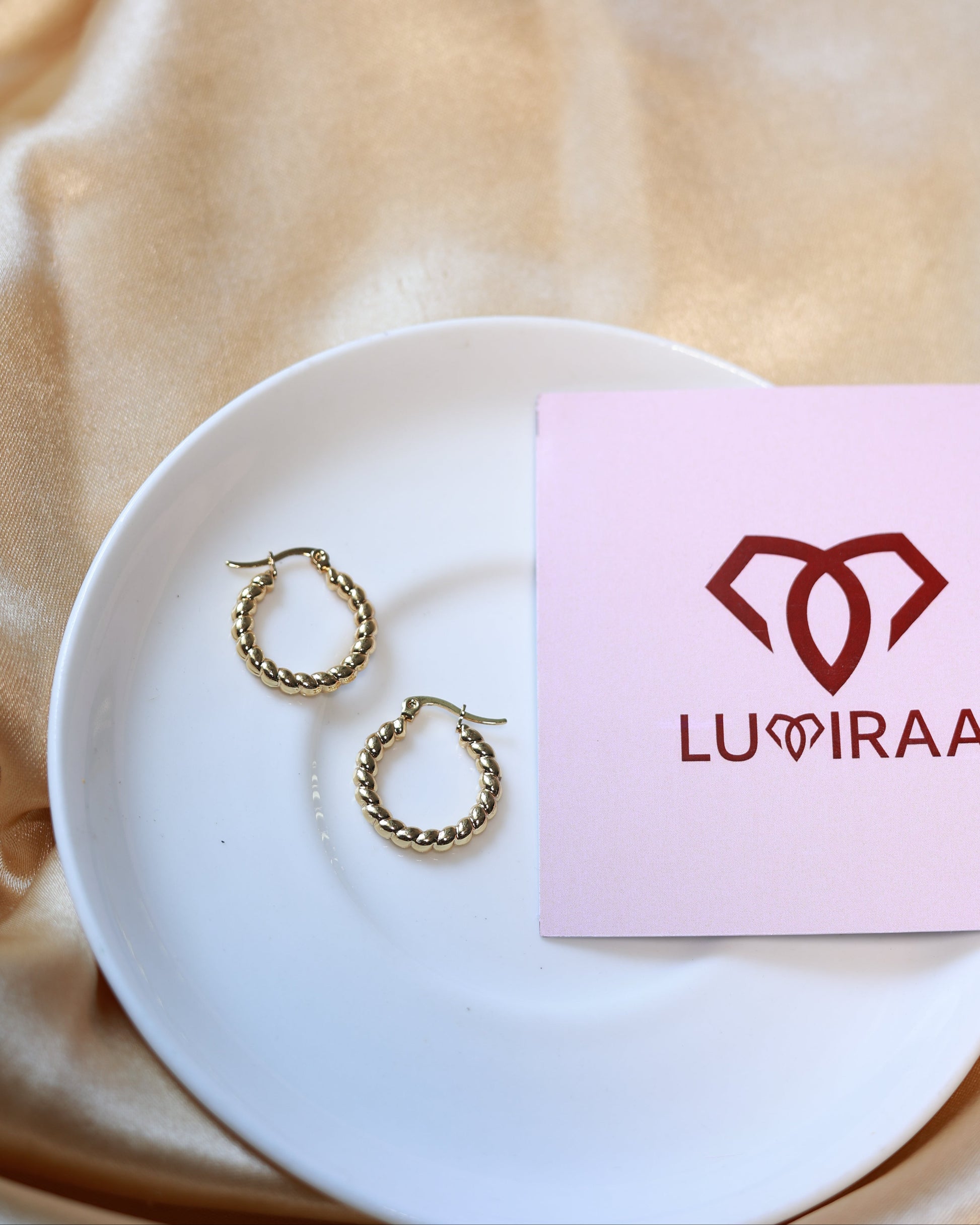 Gold hoop earrings on a white plate with a pink card featuring a logo, all on a beige fabric background.